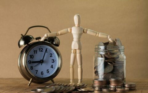 Wooden mannequin holding an alarm clock and a jar full of coins, symbolizing time and money.