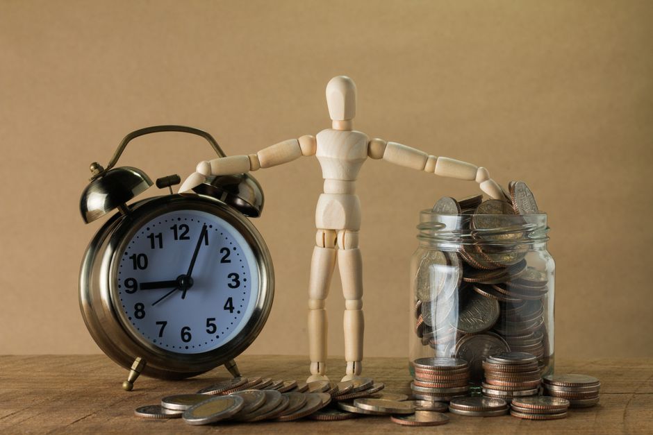 Wooden mannequin holding an alarm clock and a jar full of coins, symbolizing time and money.