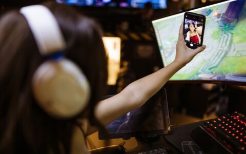 A woman wearing headphones takes a selfie with her smartphone while engaged in gaming indoors.
