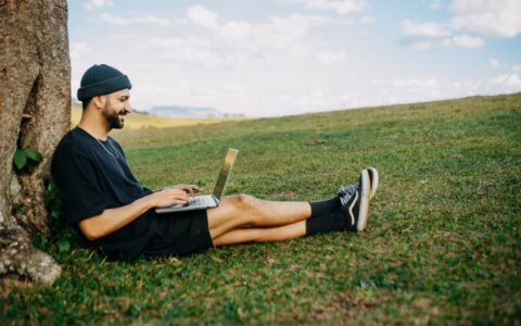 Man sitting by a tree on grass, working on a laptop and smiling. Outdoor work and relaxation.
