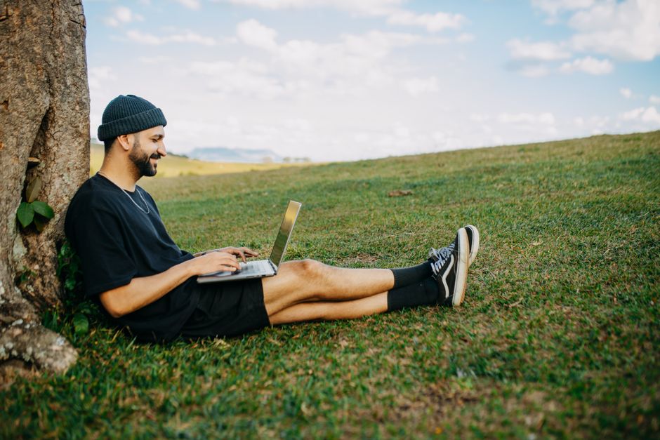Man sitting by a tree on grass, working on a laptop and smiling. Outdoor work and relaxation.