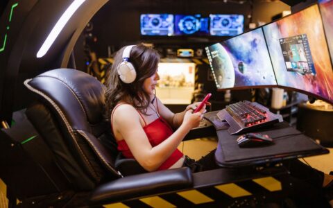 Young woman enjoying computer gaming at home with a high-tech gaming setup and smartphone.