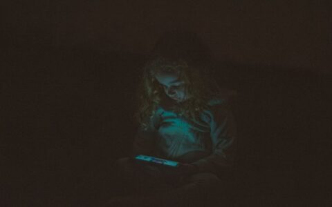 Focused little girl with long curly hair in casual clothes sitting and using smartphone while watching video in dark room