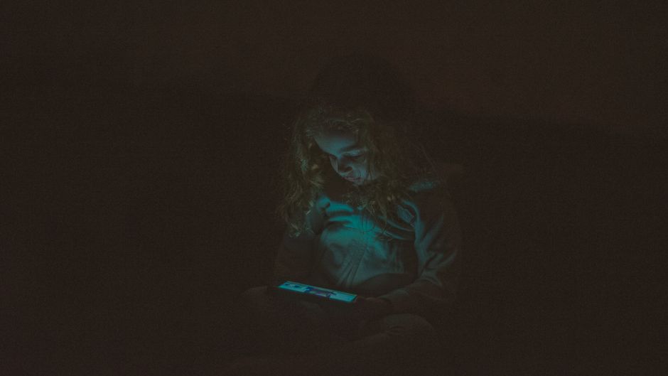 Focused  little girl with long curly hair in casual clothes sitting and using smartphone while watching video in dark room