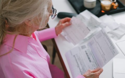 An elderly woman in glasses holds and reads important papers at a table indoors.