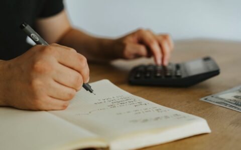 Close-up of hands writing calculations in a notebook with a calculator, focused on budgeting or financial work.
