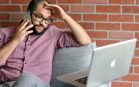 Bearded man looks frustrated while working remotely with laptop and phone.