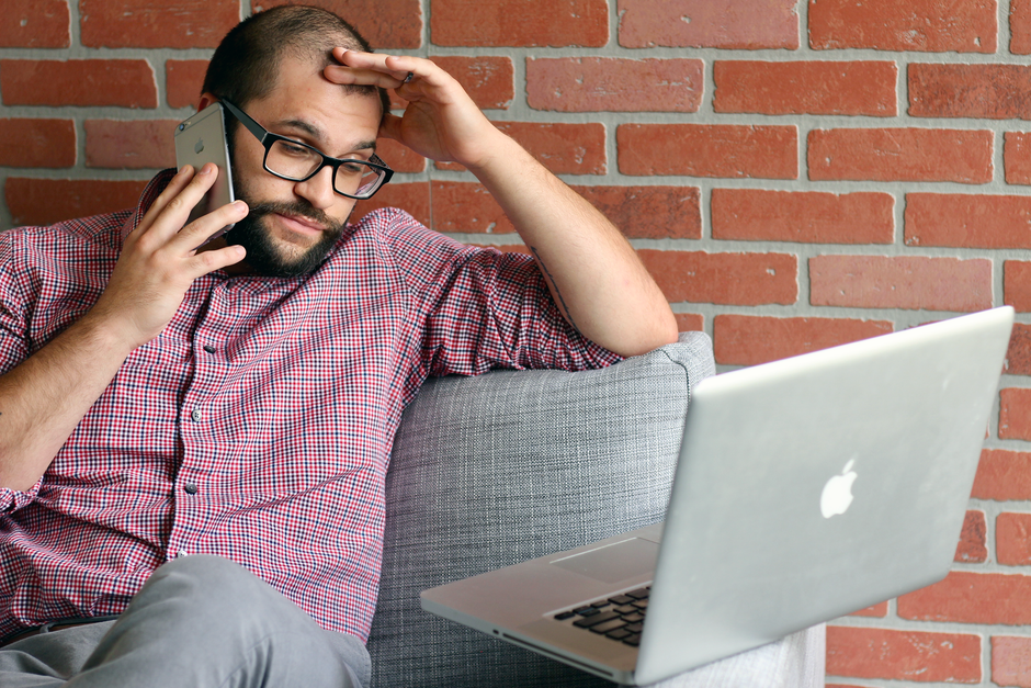 Bearded man looks frustrated while working remotely with laptop and phone.