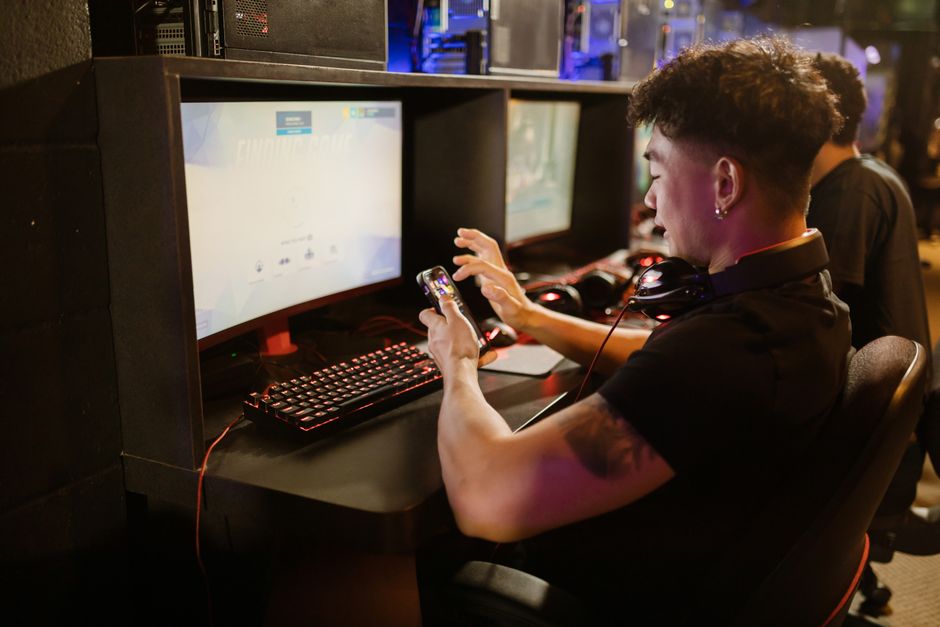 A young man is gaming using a smartphone and a PC setup with a mechanical keyboard indoors.