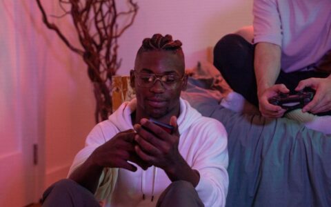 Two friends enjoying a gaming session at home, one using a mobile phone and the other a game controller.