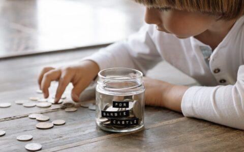 A young child collects coins in a jar labeled 'For Barbie Castle', symbolizing saving and dreams.